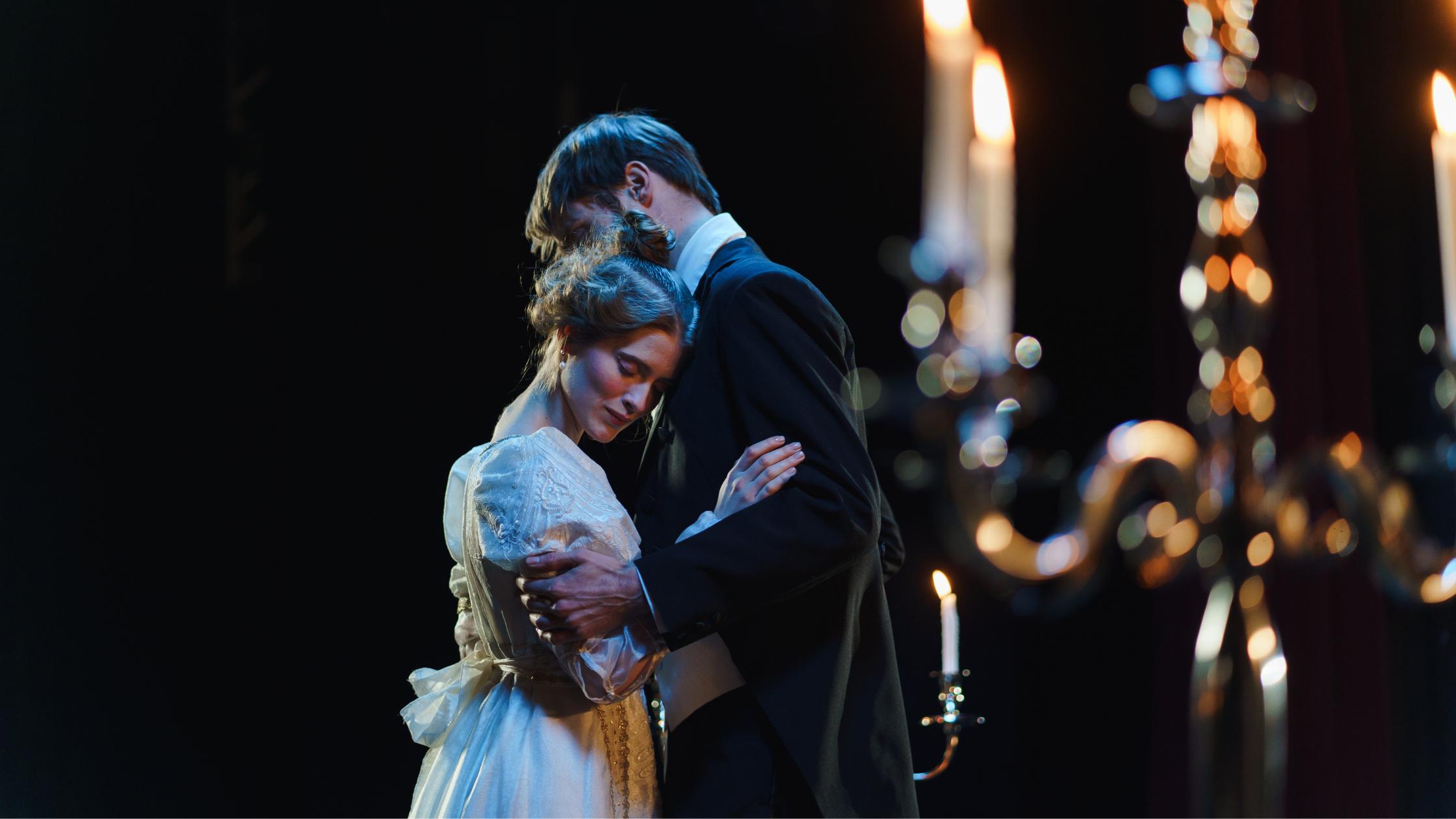 An actor and actress dressed in period garb performing an embrace on stage.